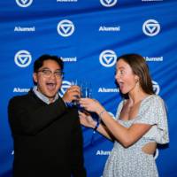 Two grads smile with champagne in front of Alumni backdrop